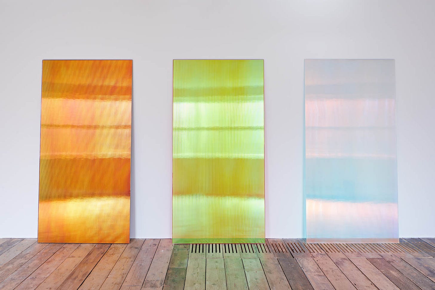 A photo showing three large rectangular panels leaning against a white wall in a gallery. From left, we have an orange panel, a light green panel, and an iridescent light blue/pink panel. It's art.