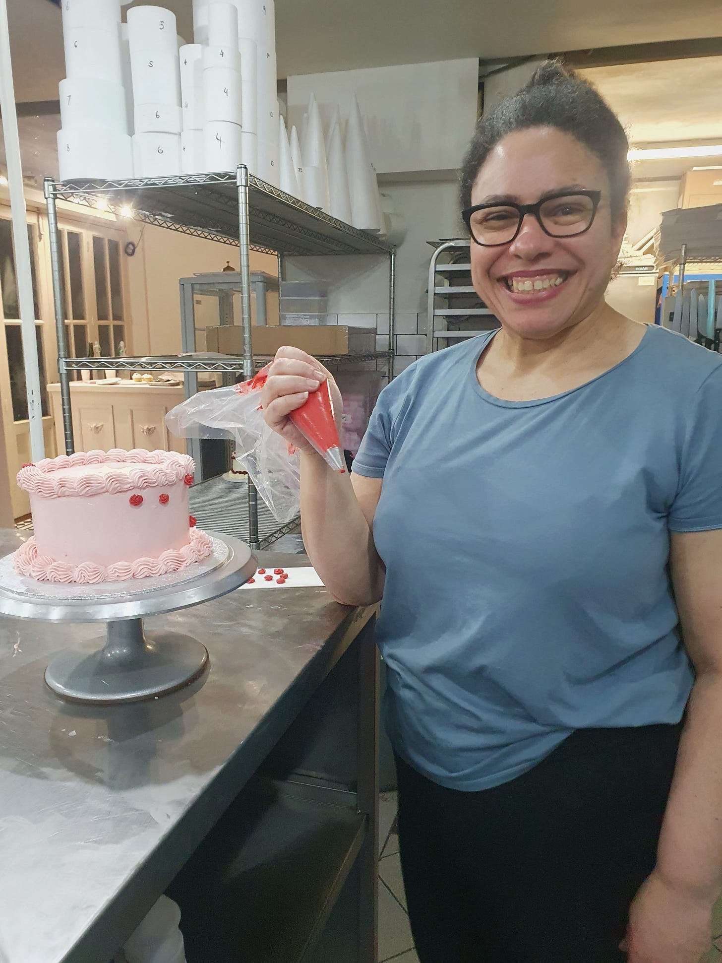 Photo of a woman holding a piping bag filled with red icing, she is looking at the camera and smiling, there is a cake covered in pink icing in front of her and she has piped some red roses onto it.