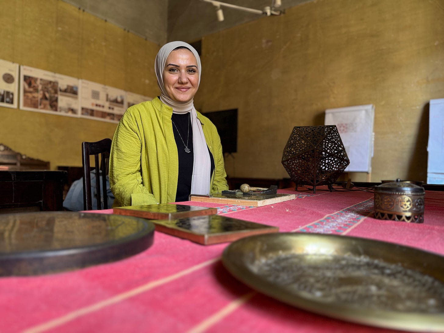 Noha El Qahwagy seated at a table displaying her handcrafted brasswork in a workshop setting.