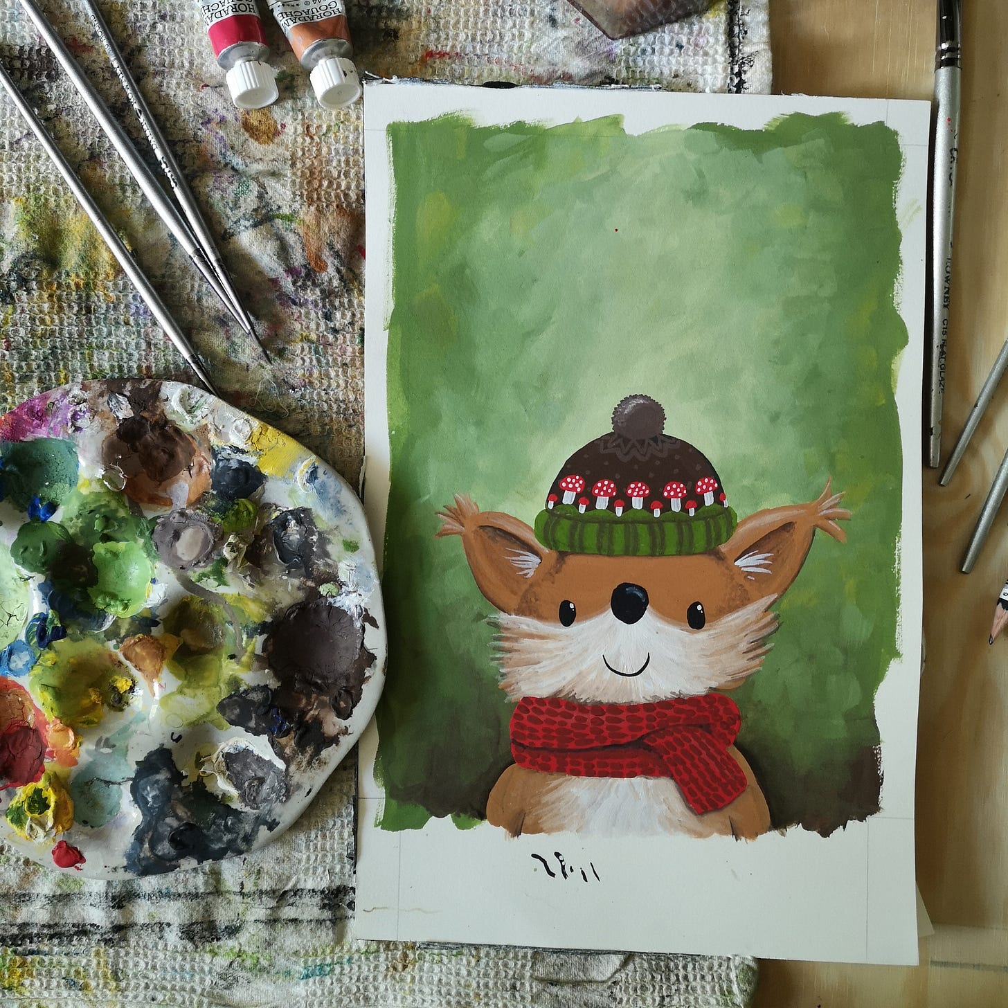 Image description: a studio table shown from above, with paint palette, brushes, tubes of gouache and a painting-in-progress all lying on top of a very dirty tea towel. The painting shows the head and shoulders of a supremely cute red squirrel who is wearing a red woollen scarf and a hat with green brim, dark brown background with mid bron spots, a parade of red and white mushrooms all around, and a pom pom on top. The background of the painting is green.