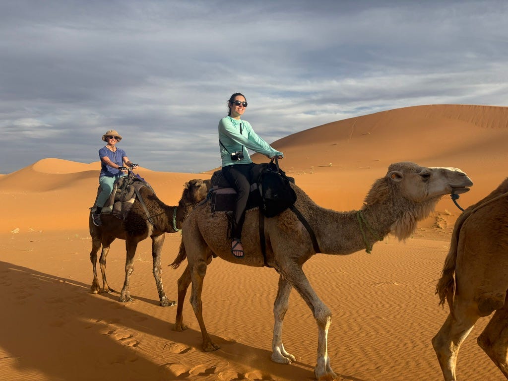 Two women wearing sunglasses and blue shirts ride camels with rippling, orange sand dunes in the background. Two women wearing sunglasses and blue shirts ride camels with rippling, orange sand dunes in the background.