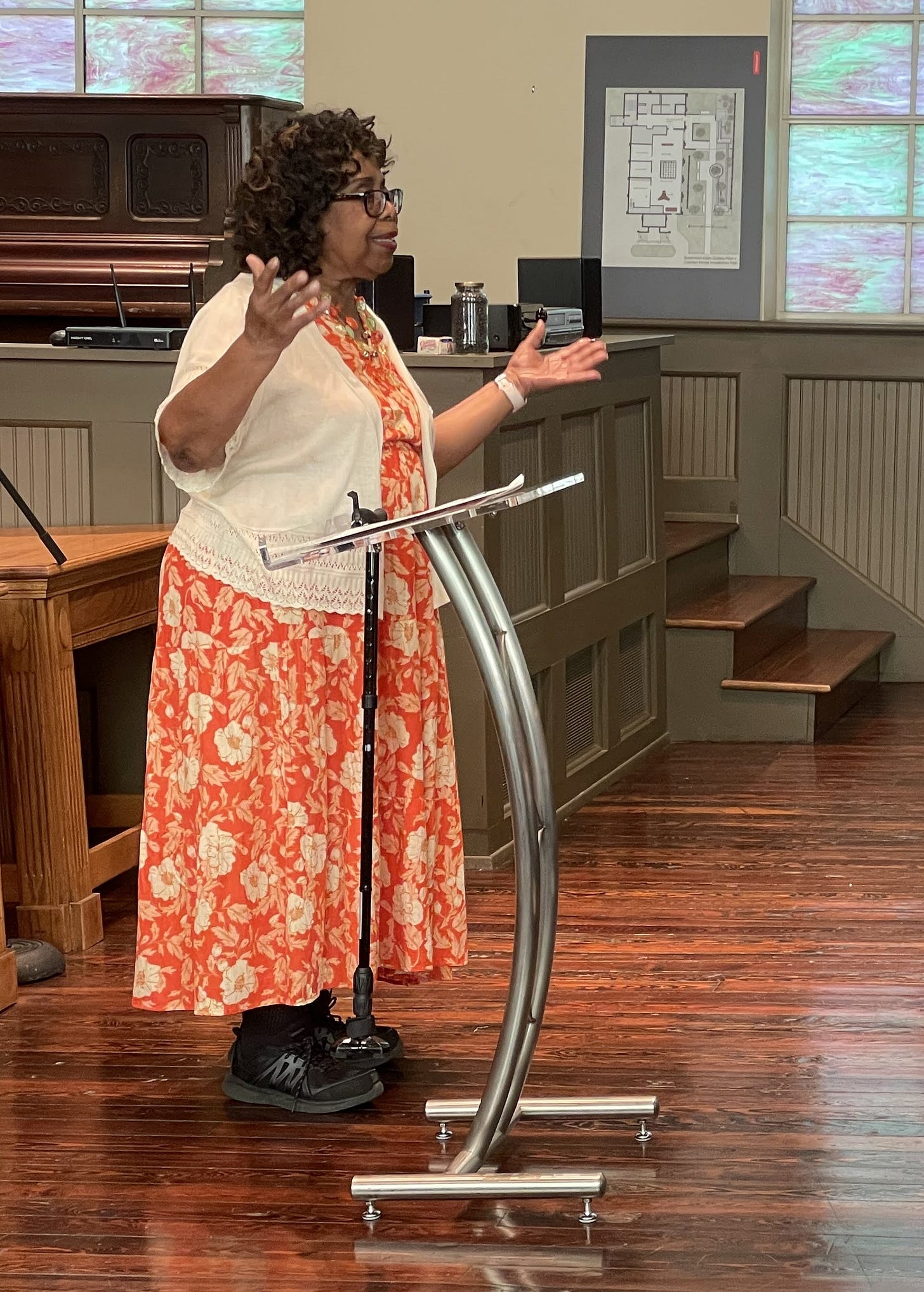 Black woman with curly hair and glasses wearing an orange print dress and cream sweater stands at a podium, her arms raised as she speaks