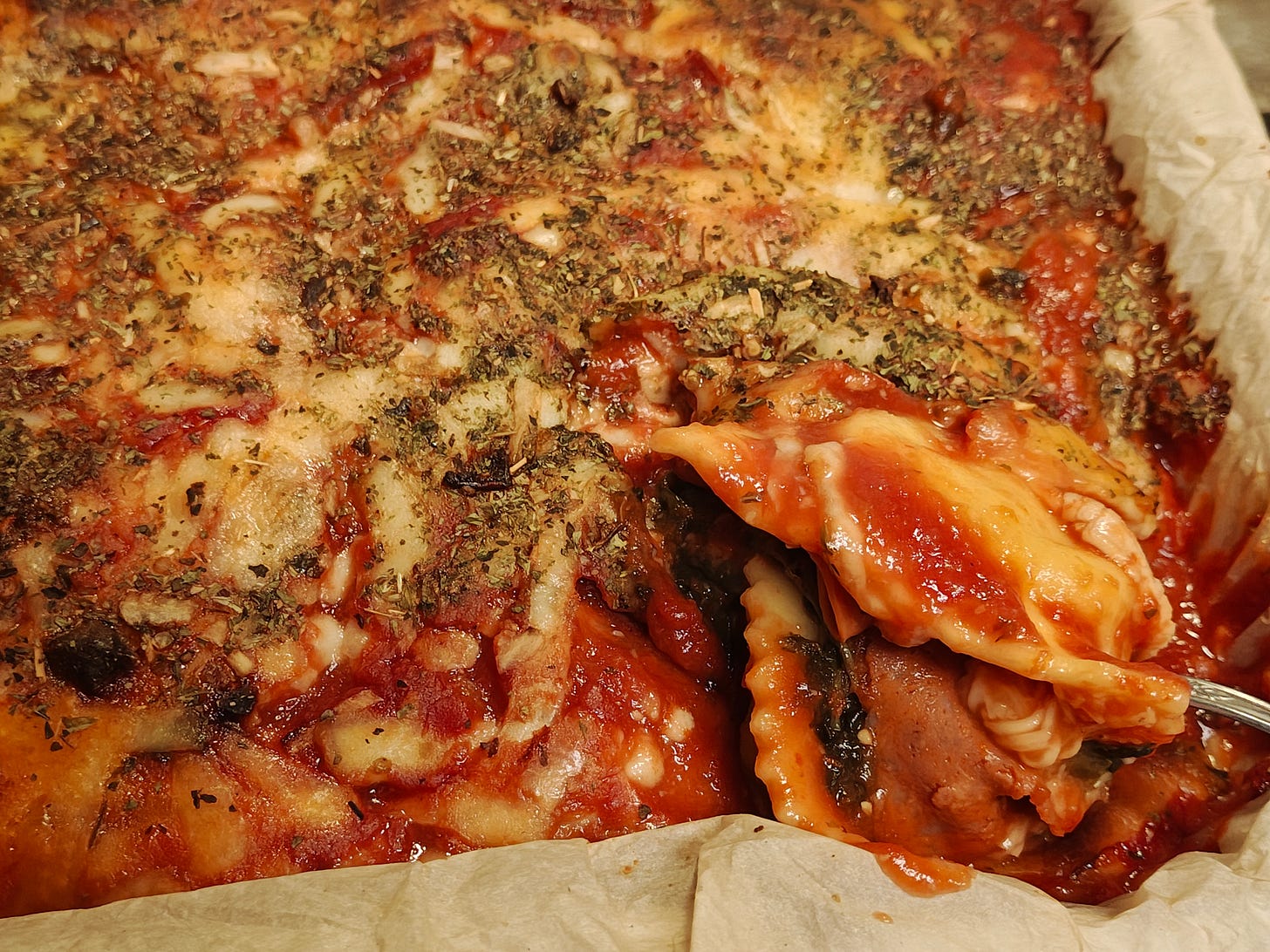 Close-up of a pepperoni ravioli casserole covered with marinara, melted browned cheese, and dried herbs. A fork lifts one saucy ravioli from the dish, revealing layers of spinach, pepperoni, and cheese beneath the surface.
