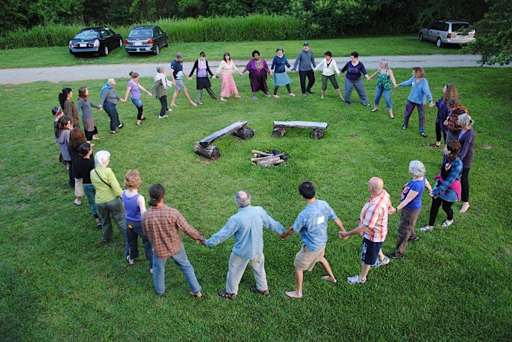 a large multigenerational circle of people hold hands around a fire pit on a lush green lawn in the country