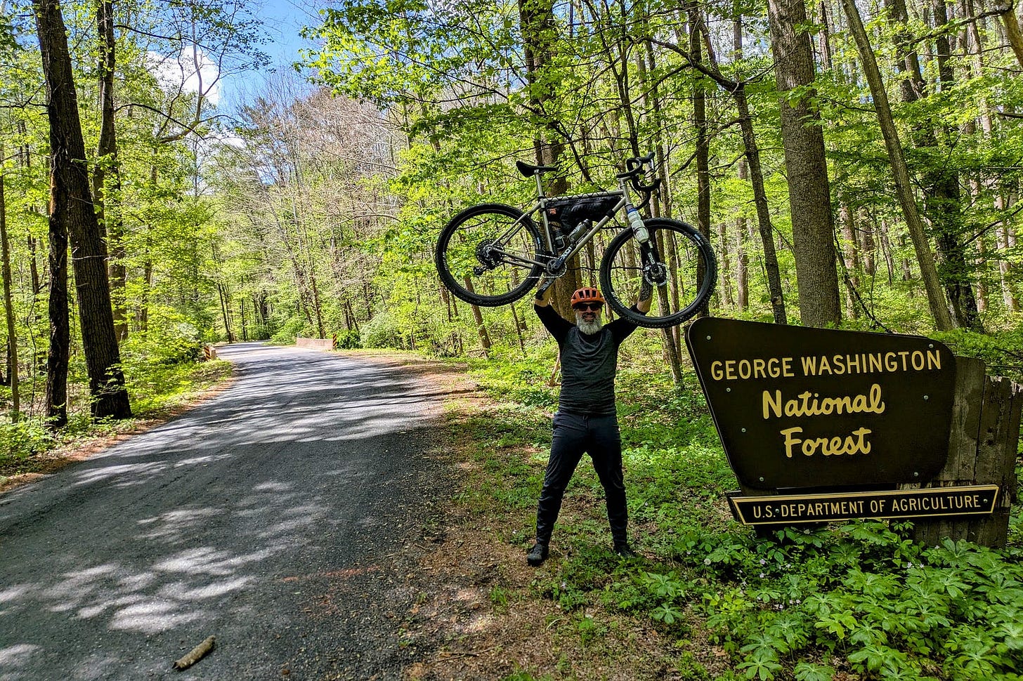 The author stands beside a road sign for George Washington National Forest, holding a gravel bike overhead. He wears a helmet and cycling gear, surrounded by tall trees and greenery along a quiet forest road. The author stands beside a road sign for George Washington National Forest, holding a gravel bike overhead. He wears a helmet and cycling gear, surrounded by tall trees and greenery along a quiet forest road.