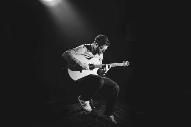 >grayscale photo of man playing guitar on stage >grayscale photo of man playing guitar on stage