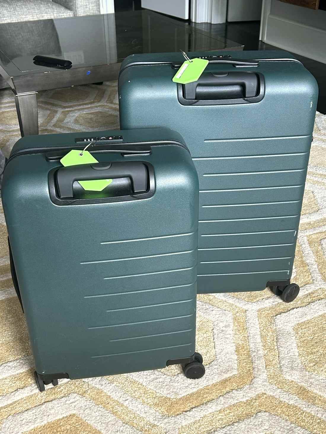 Two dark green hard-shell Quince suitcases on a patterned rug.