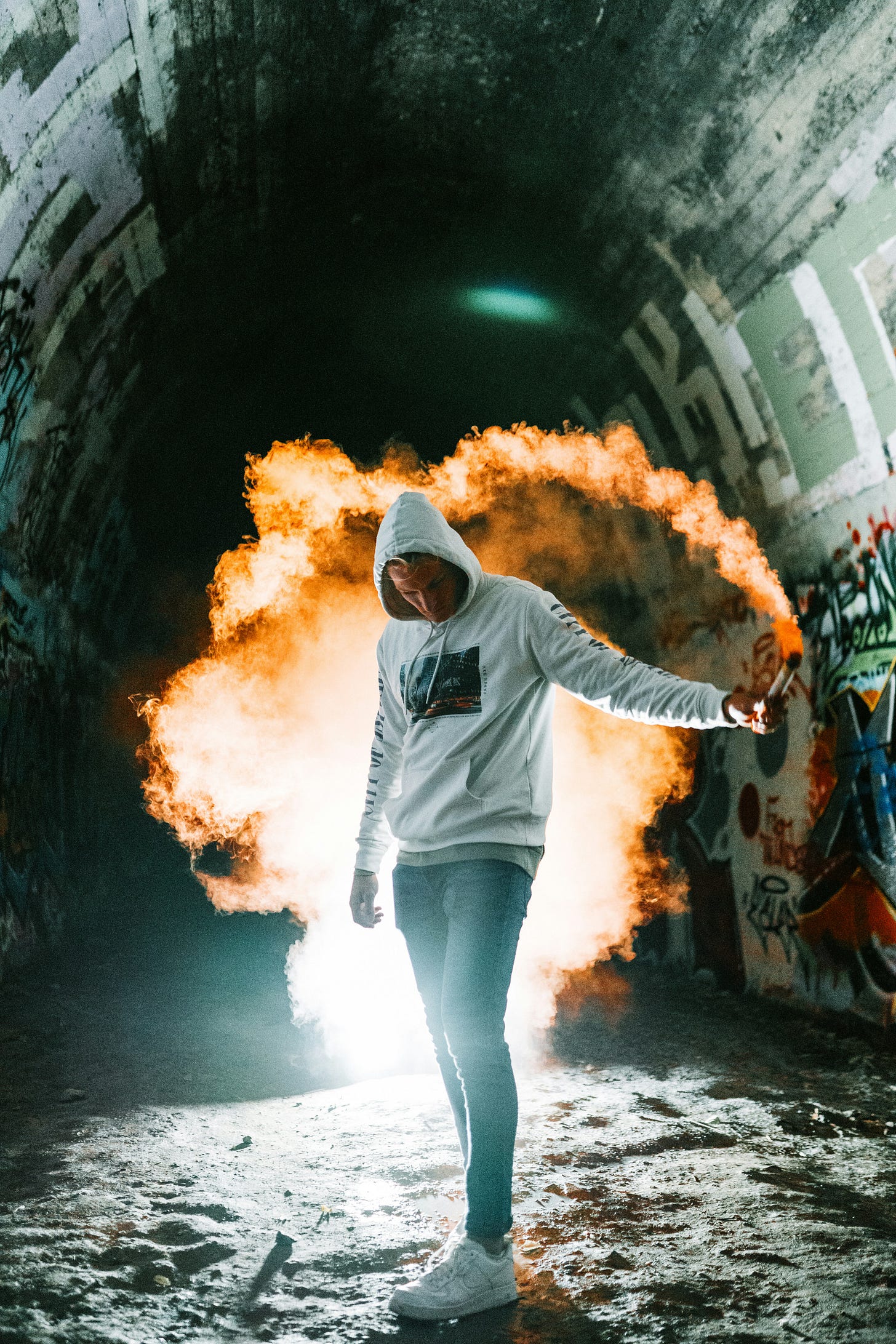 A person wearing a white hoodie and jeans holding a lit smoke flare in an urban graffiti-covered tunnel, with dramatic orange smoke and bright light creating a rebellious and bold atmosphere.