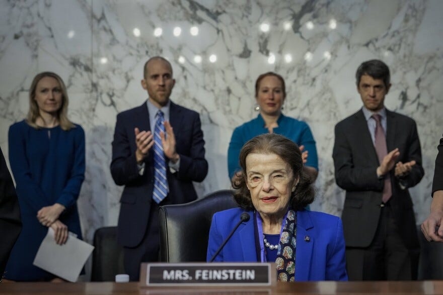 An elderly woman wears a cobalt blue jacket with pearls as she sits at a dais where the name plate reads: Mrs. Feinstein. People stand behind her, some clapping. An elderly woman wears a cobalt blue jacket with pearls as she sits at a dais where the name plate reads: Mrs. Feinstein. People stand behind her, some clapping.