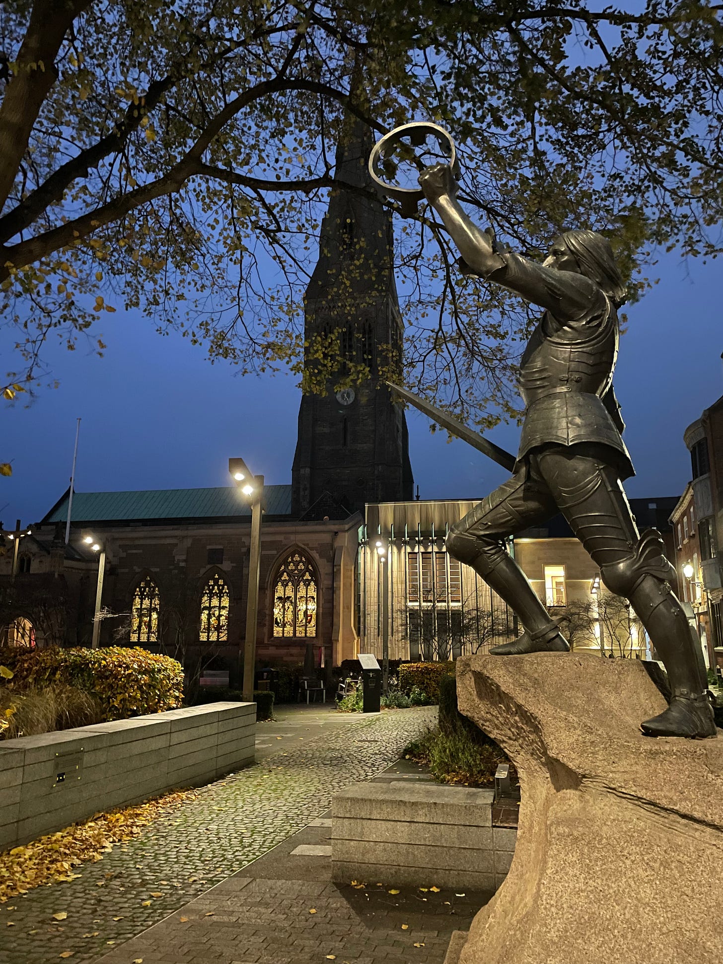 A statue of Richard III on a plinth, holding a crown aloft in one hand and a sword in the other. It's dusk.
