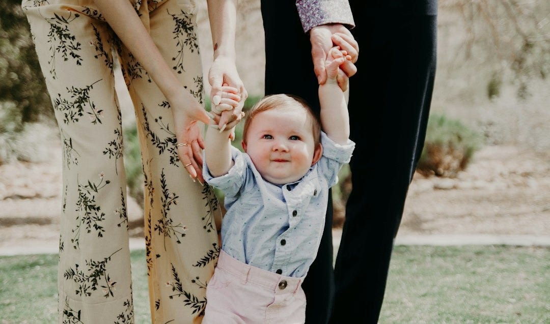 man and woman holding hand of toddler walking on grass field
