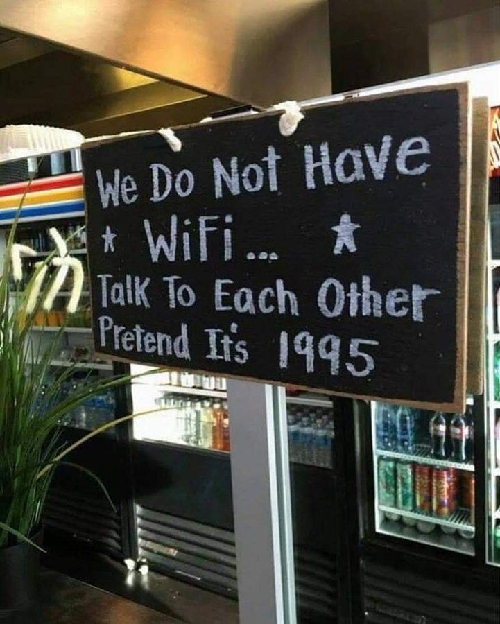 The image shows a black chalkboard sign hanging in what appears to be a cafe, with a well-lit refrigerator stocked with drinks and a plant visible in the background. The sign, written in white chalk, humorously states, "We Do Not Have * WiFi... * Talk To Each Other Pretend It\'s 1995," encouraging social interaction. The post text from Varsha Singh (@varshaparmar06) labels it as part of a "wholesome thread" promising happiness, enhancing the positive, nostalgic vibe. No platform watermarks are present, and the composition is clear and engaging, reflecting a playful meme about disconnecting from technology.