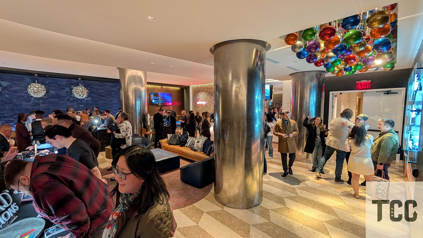 A wide-angle view of the crowd at The Indies event, with guests gathered around watch display tables. A cluster of colorful balloon-like ceiling lights adds a playful contrast to the otherwise sleek, modern space.