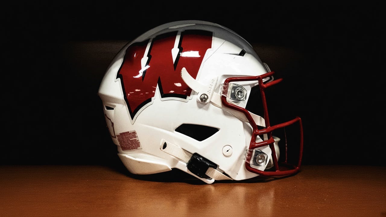 Close-up of a white Wisconsin Badgers football helmet with a red Motion W logo and red facemask against a dark background. Close-up of a white Wisconsin Badgers football helmet with a red Motion W logo and red facemask against a dark background.