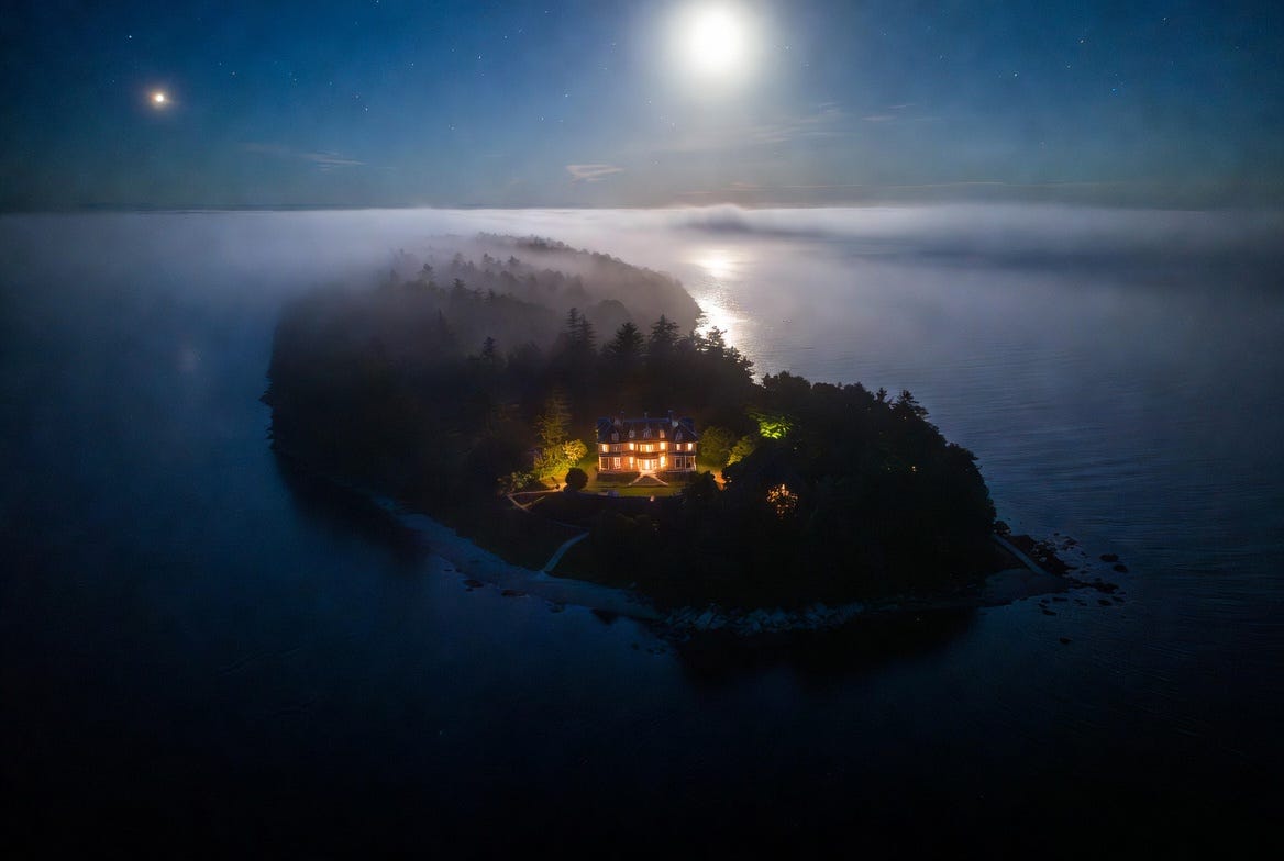 Aerial night view of Jekyll Island off the Georgia coast, fog over dark water, a single illuminated Victorian clubhouse visible through dense trees
