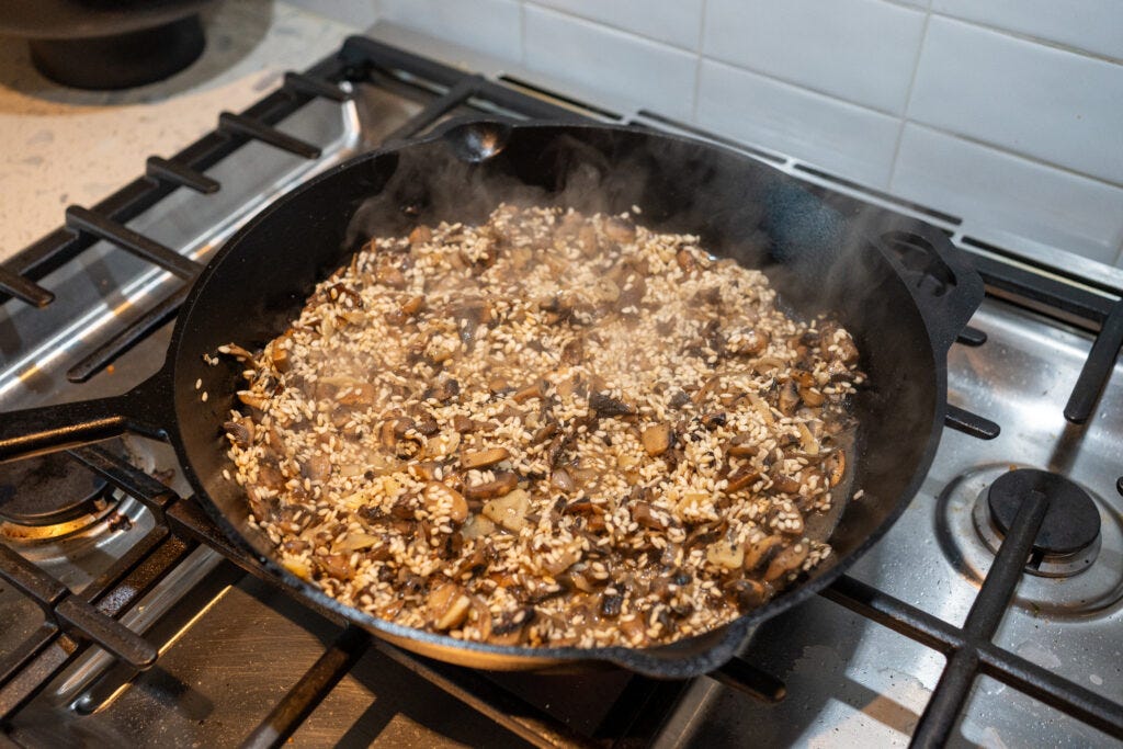 Raw Arborio rice grains being stirred and toasted with a savory mushroom and onion base in a cast iron skillet.