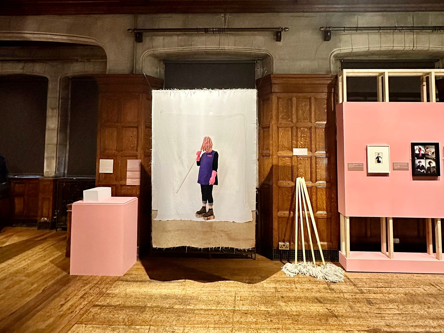 Photo of a large photographic mural in an expansive hall. The photo shows a cleaner in uniform holding a mop up to their face and covering it. Photo of a large photographic mural in an expansive hall. The photo shows a cleaner in uniform holding a mop up to their face and covering it.