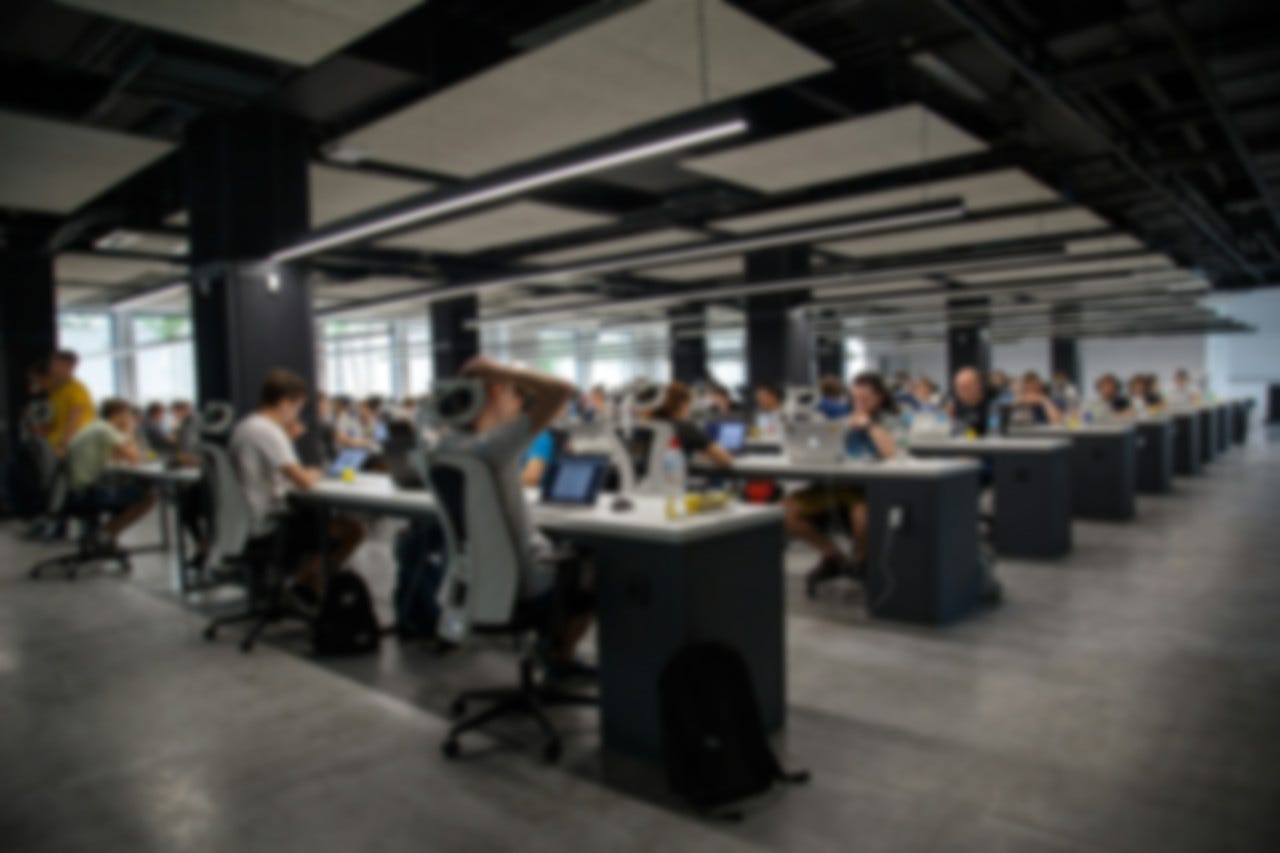 Large room with numerous desks with people hunched over their laptops