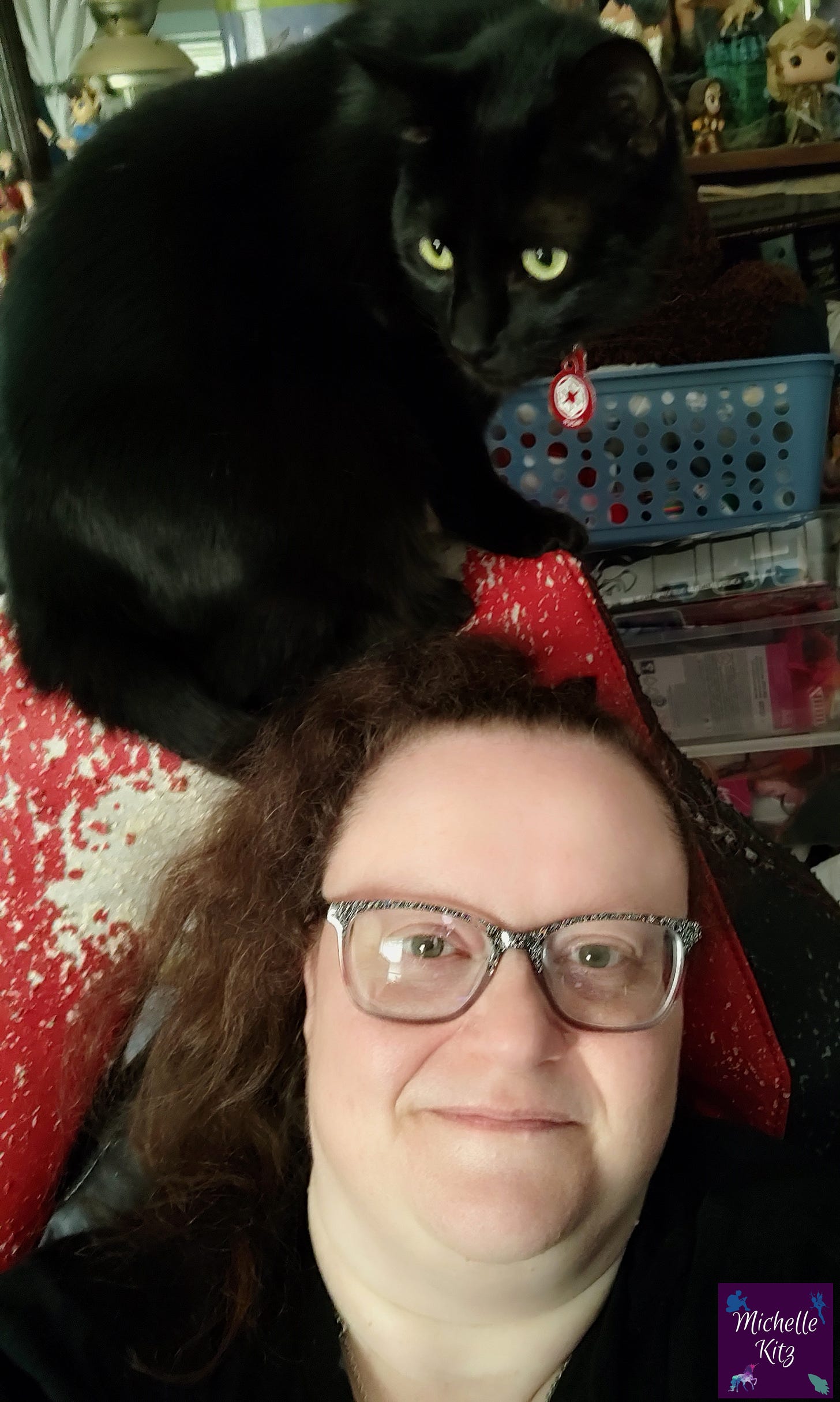 Author Michelle Kitz sitting in a red and black desk chair with her black cat, Vader perched on the back of the chair.