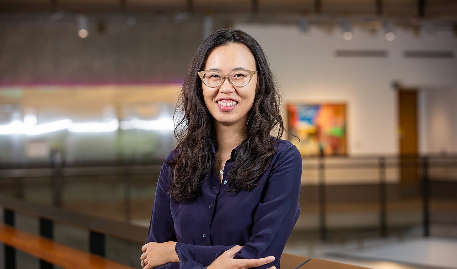 a photograph of a woman with brown wavy hair below her shoulders in a purple/navy button up shirt. Her arms are folded and she has beige retro-style glasses. She’s smiling and appears to be in her thirties. 