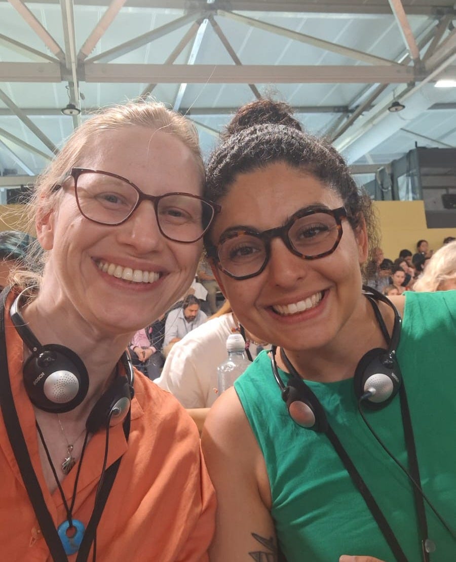 Foto selfie. Eu, de cabelos presos mas bagunçados, e Mari. Ambas com fones de tradução simultânea sorrindo apesar do cansaço.