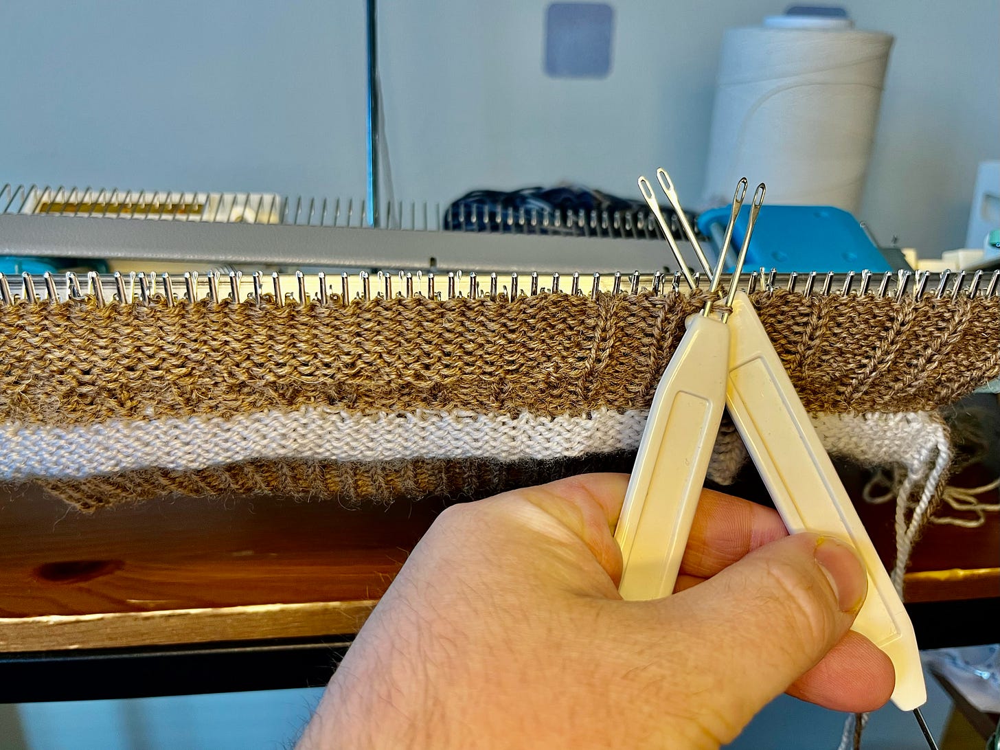 A hand holds two cream-coloured transfer tools over the needle bed of a knitting machine. A section of striped brown and white knitted fabric hangs from the needles. The tools are being used to move individual stitches, as required for forming cables.