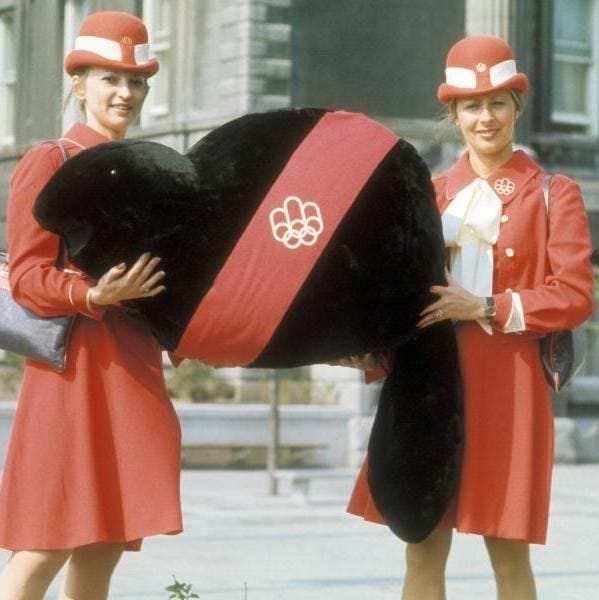 This color photograph depicts two young women in matching red uniforms—consisting of knee-length dresses, white-collared blouses with bow ties, pillbox hats featuring white bands and Olympic emblems, and small shoulder bags—standing outdoors on a paved plaza in front of a grand stone building with classical columns and arched windows, suggestive of a historic urban setting.

They smile warmly at the camera while collaboratively holding a large, oversized black plush beaver mascot named Amik, characterized by its rounded body, flat tail, small ears, and beady eyes. The beaver wears a wide red sash across its midsection emblazoned with the white interlocking Olympic rings. The scene is set under a clear sky with subtle greenery and steps in the background, conveying a promotional or celebratory atmosphere.