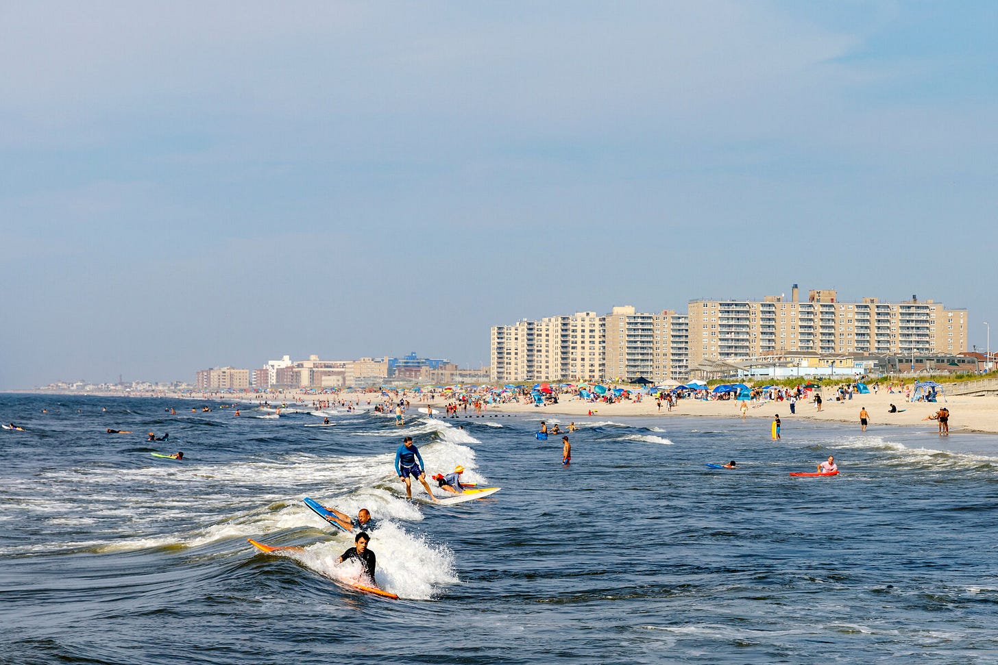 A picture of surfers on the sea, against the backdrop of the sand and buildings of Rockaway Beach