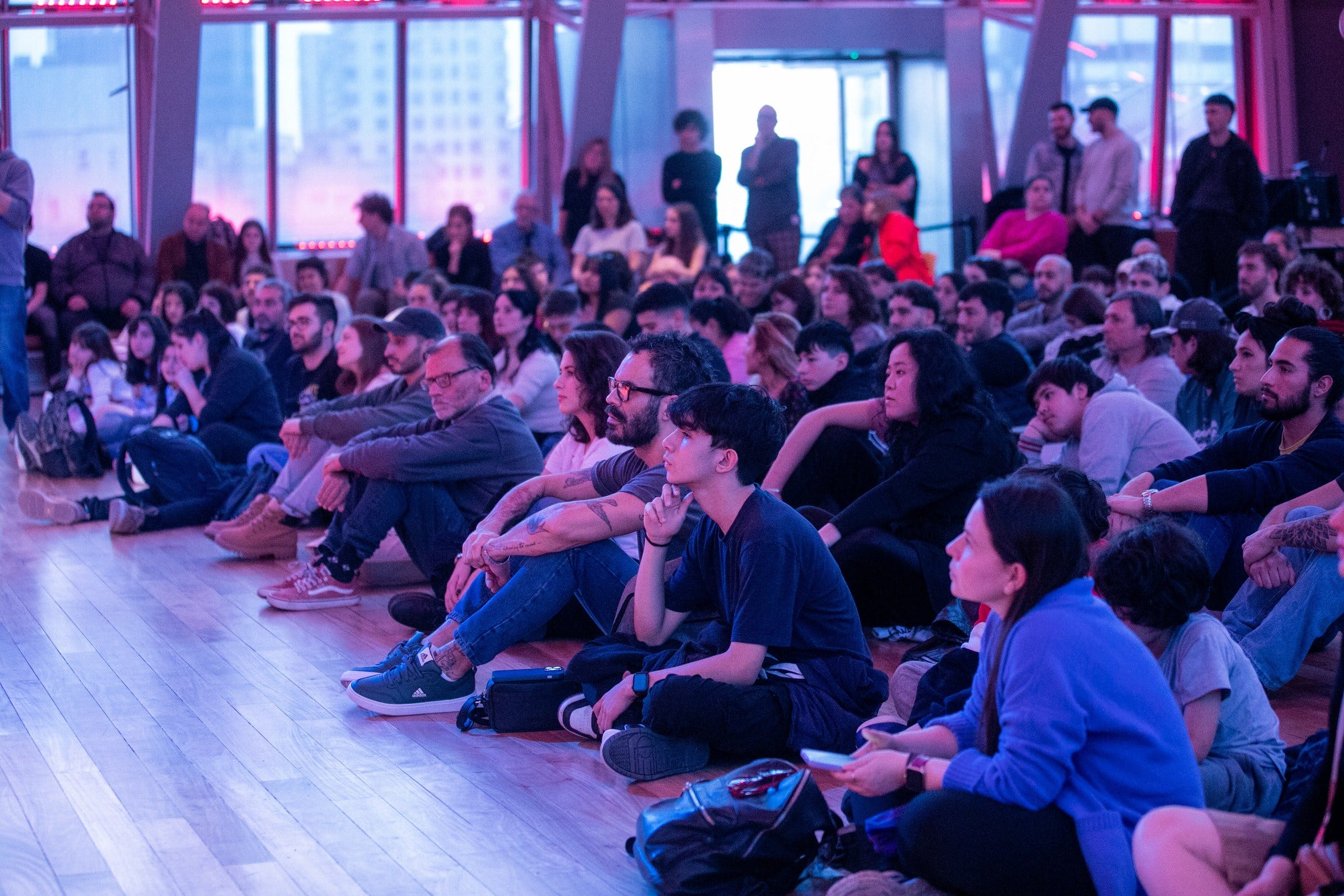 A wide view of the audience seated on the floor of the Palacio Libertad hall, washed in pink and blue light, during Resonancias Paralelas A wide view of the audience seated on the floor of the Palacio Libertad hall, washed in pink and blue light, during Resonancias Paralelas