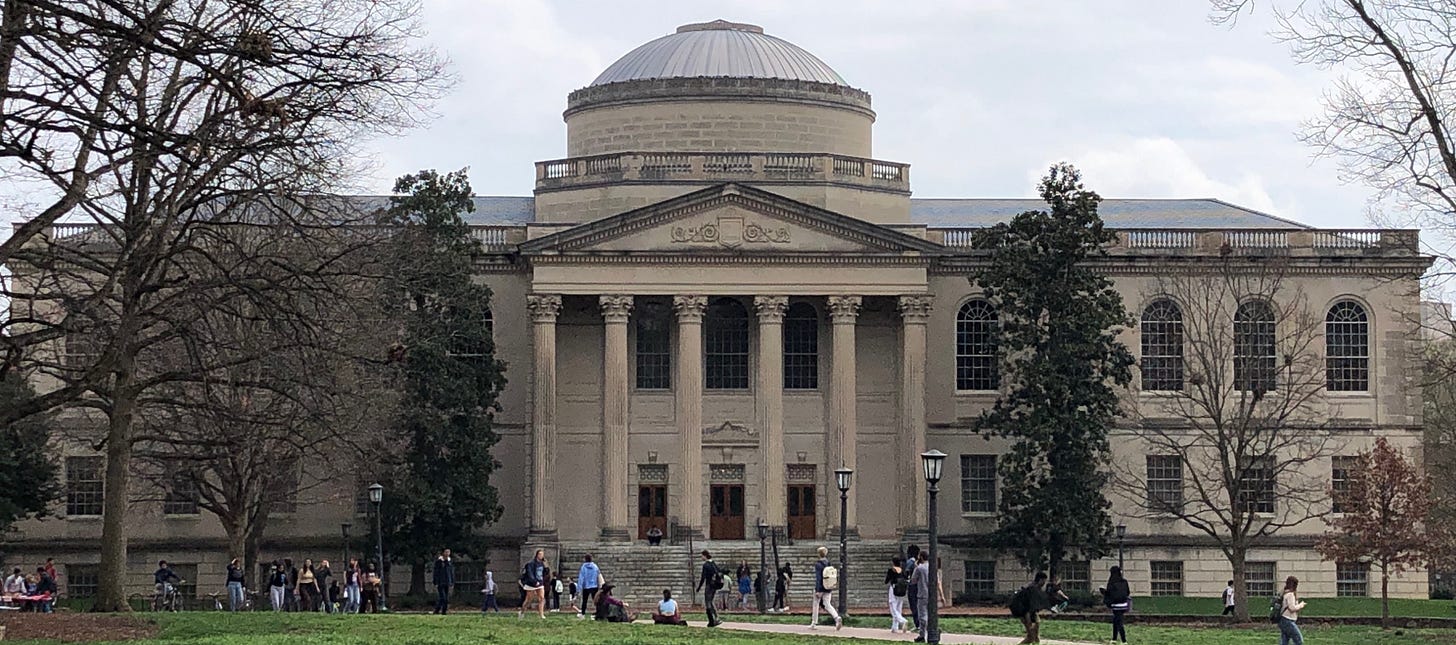 Long, rectangular building fronted with columns and domed ceiling. Lawn with students and a few trees.