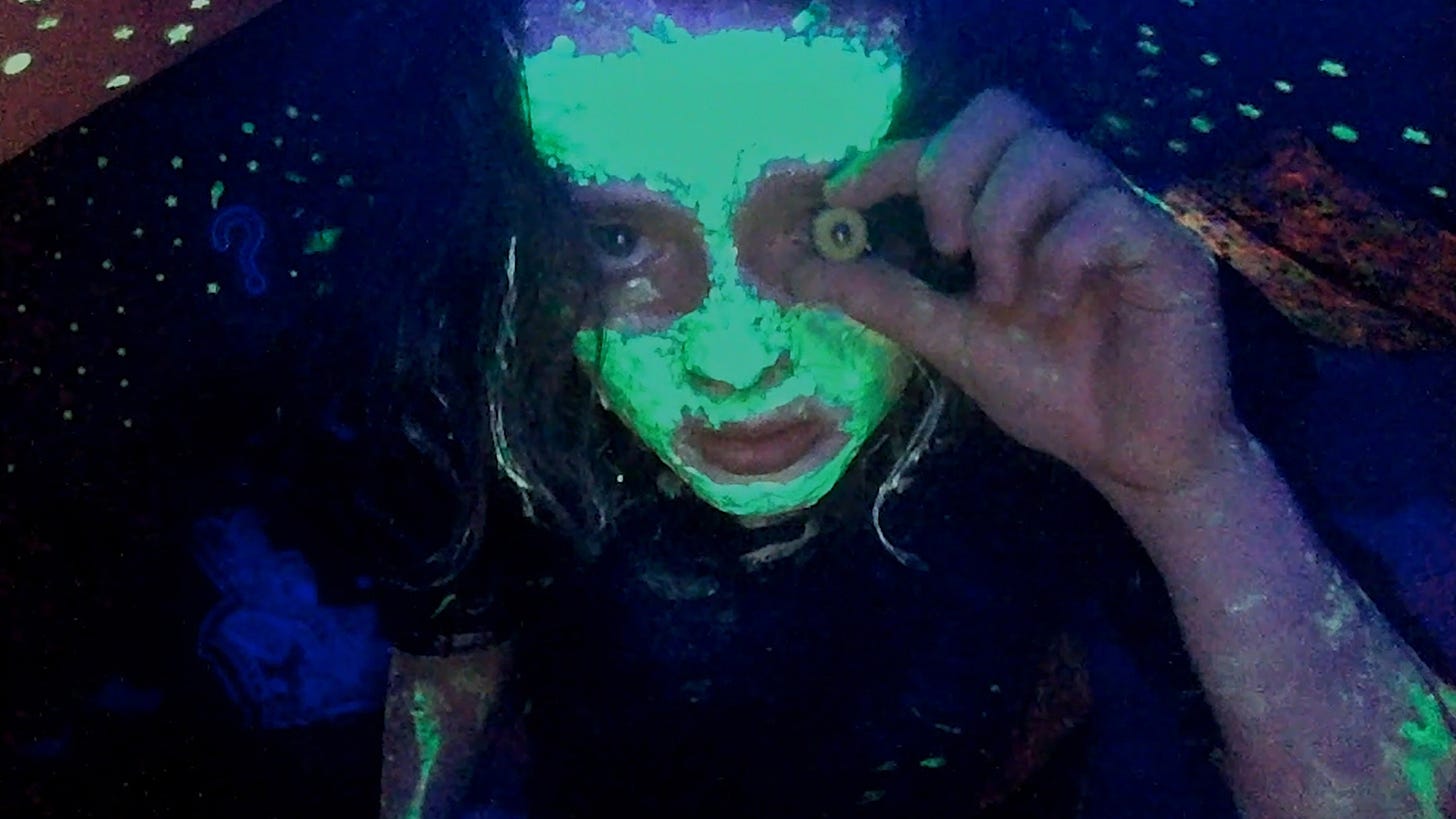 Character named Casey, covered in glowing face paint, stares into a webcam, holding a button to her eye. Character named Casey, covered in glowing face paint, stares into a webcam, holding a button to her eye.