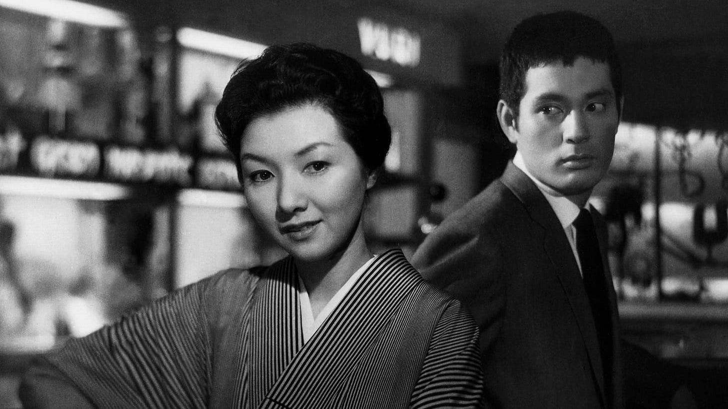 When a Woman Ascends the Stairs | Still features Hideko Takamine looking directly at the camera with a smile, while Tatsuya Nakadai watches from behind.