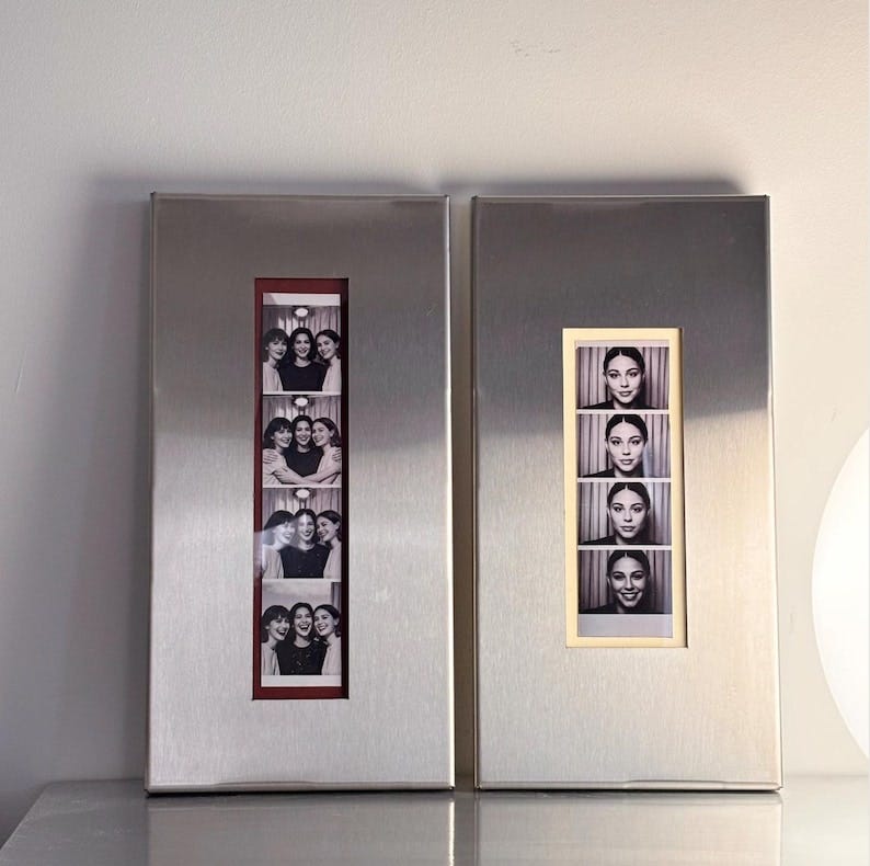 May include: Two rectangular, brushed metal photo frames. One frame displays a series of black and white photo booth images of three women. The other frame shows a series of black and white photo booth images of a woman.