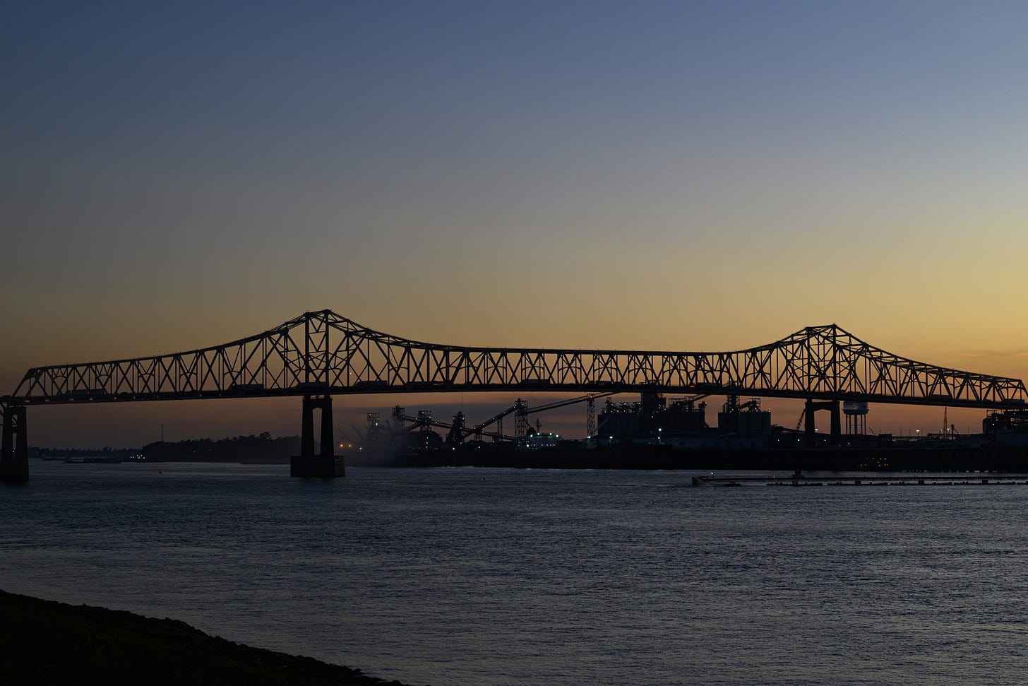 Silhouette of a bridge at dusk, spanning over a body of water, with industrial structures and soft sunset colors in the background. Silhouette of a bridge at dusk, spanning over a body of water, with industrial structures and soft sunset colors in the background.