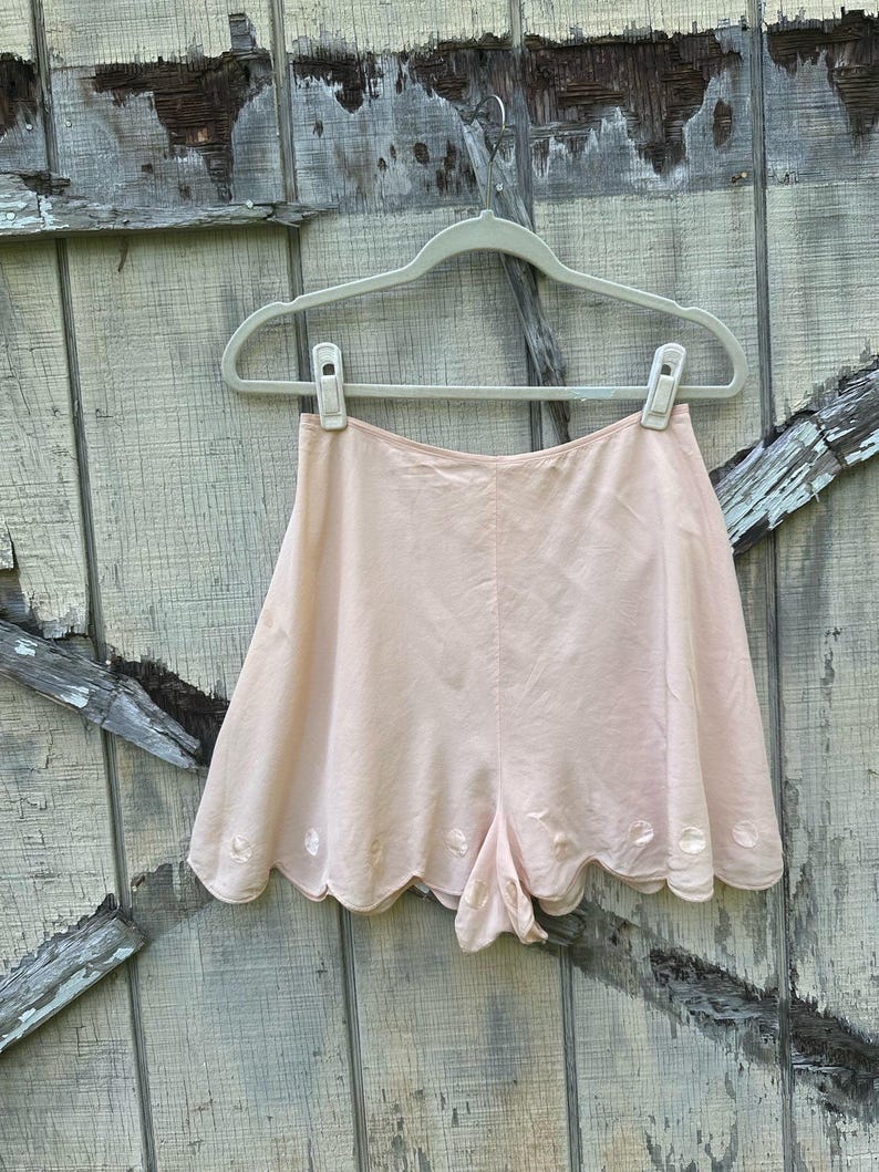 May include: A pair of light pink shorts with scalloped edges and embroidered white flowers. The shorts are hanging on a white plastic hanger against a wooden background.