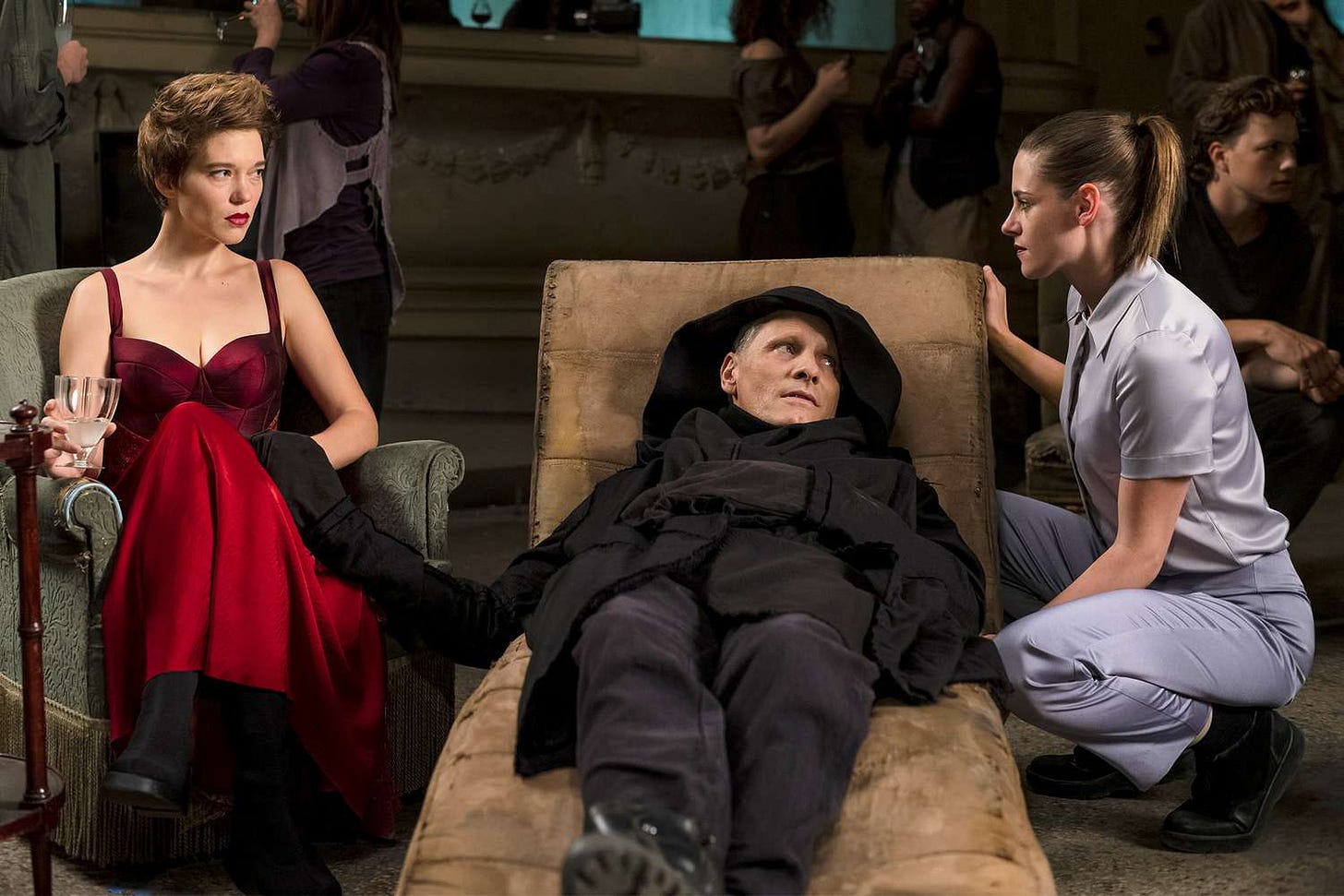 Saul Tenser (a silver-haired man clad in heavy black clothes with a hood) reclines in a divan in evident exhaustion. He's flanked by a glaring woman in a red satin party dress (Lea Seydoux) and kneeling brunette in scrubs (Kristen Stewart). Saul Tenser (a silver-haired man clad in heavy black clothes with a hood) reclines in a divan in evident exhaustion. He's flanked by a glaring woman in a red satin party dress (Lea Seydoux) and kneeling brunette in scrubs (Kristen Stewart).