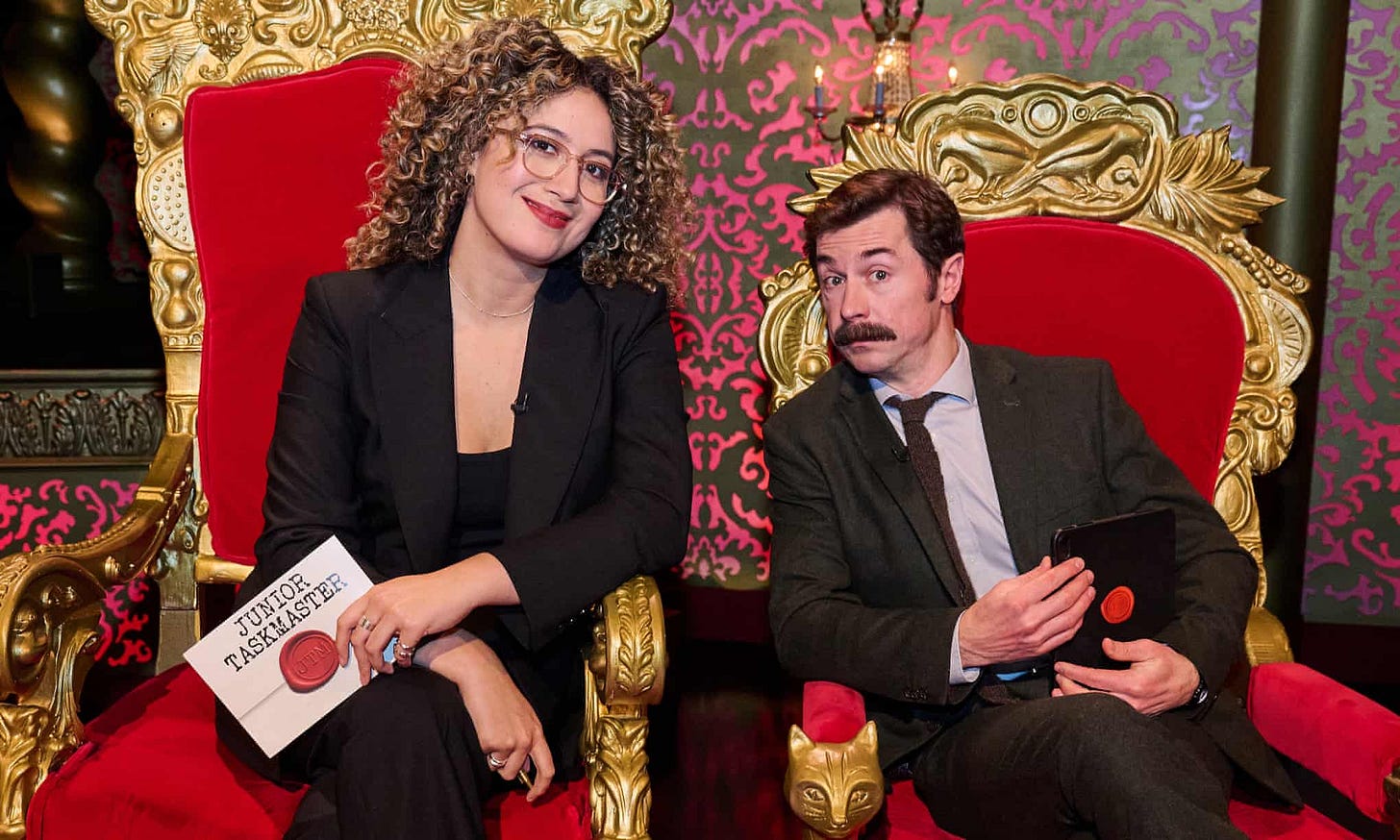 Junior Taskmaster hosts Rose Matafeo and Mike Wozniak sitting in plush red velvet thrones on the Taskmaster stage. Rose has a task card in her hand.