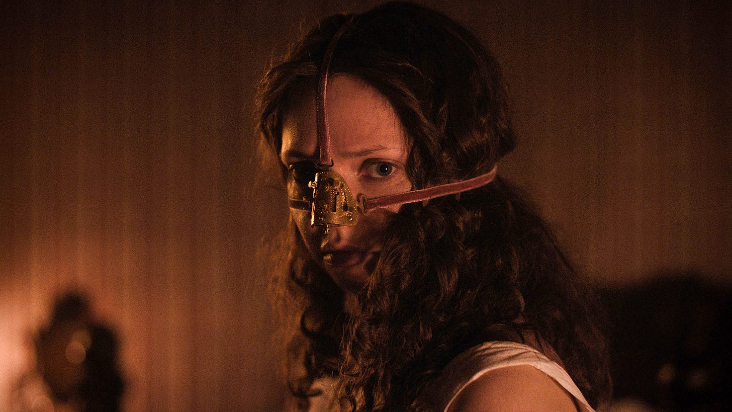 Esta imagem mostra a atriz Lea Myren como a personagem Elvira no filme de terror norueguês de 2025, A Meia-Irmã Feia (The Ugly Stepsister).  Na cena, Elvira aparece usando um dispositivo metálico preso ao nariz e ao rosto por tiras, sugerindo um dos "procedimentos cosméticos angustiantes" retratados no filme. A iluminação é sombria e em tons quentes, destacando seu olhar intenso e seu cabelo escuro e cacheado.