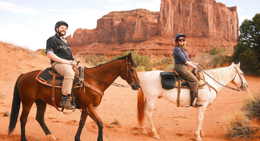 Two people sat on top of brown and white horses, with the orange rocky Monument Valley in the background