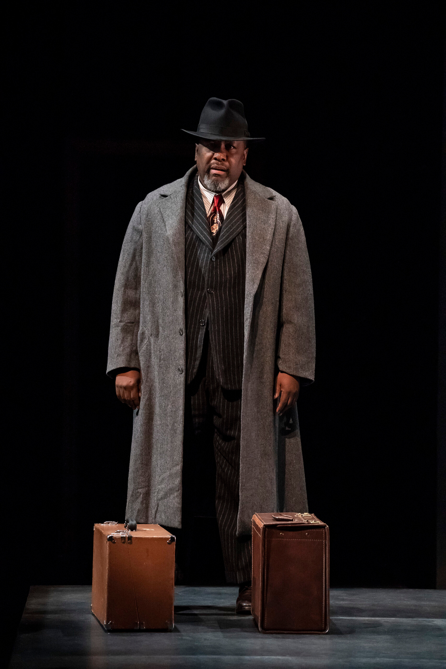 Wendell Pierce as Willy Loman in Death of a Salesman, standing alone onstage in a long overcoat and hat under dark theatrical lighting.