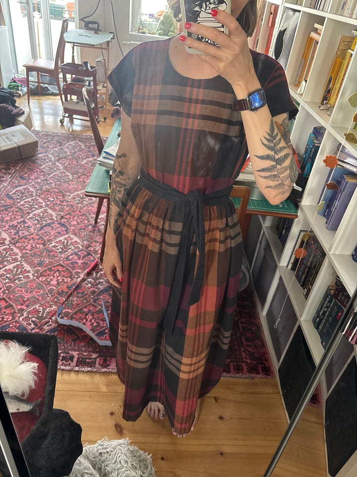 White woman with fern tattoos on her arms wears a plaid print dress with gathered waist and short sleeves in brown, cream, raspberry and pale orange. Picture on left has a loose waist with gathering free, on the right the waist is cinched in with a fabric tie belt.