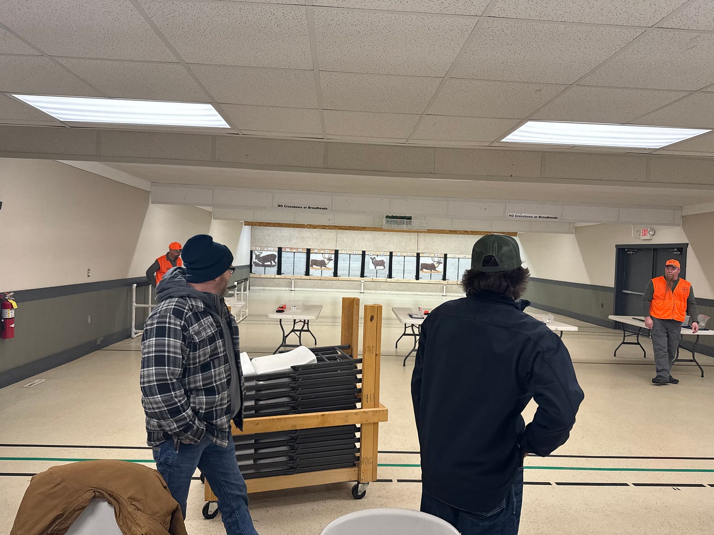 A photo of an indoor archery shooting range, a wide room with a beige linoleum floor and white drop ceiling with fluorescent lights. Against the back wall are several archery targets representing deer and elk. Lined up midground are folding tables, one oriented toward each target. Two instructors in orange vests and hats are each at a table, with others soon to take their places. In the foreground is a rolling rack of folding chairs and two dudes in flannel and denim looking back toward the targets. 