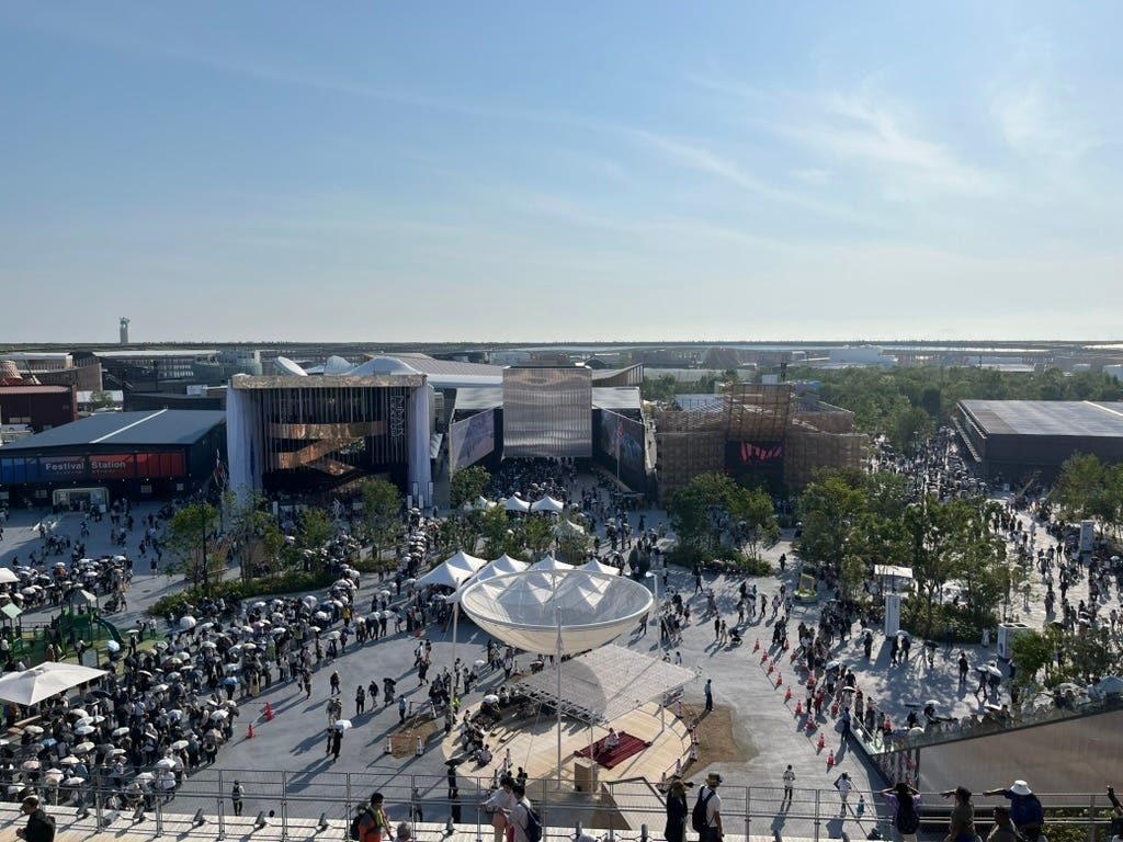 5 large buildings, from left to right: a dark, flat, boxy building labeled "Festival Station"; a taller, rectangular building with vertical hanging lines labeled "France"; a shiny, angular building with an American flag in front; a polygonal building made of wood; and a black, flat, boxy building. In front of the buildings is a plaza with many people, and there is a small performance space near the front of the plaza, with a musician performing for a small crowd.