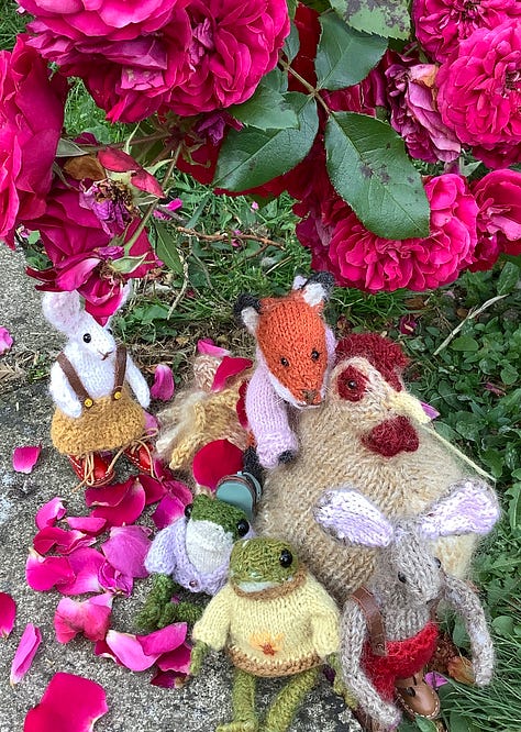 Knitted toys under a red rose bush surrounded by fallen petals