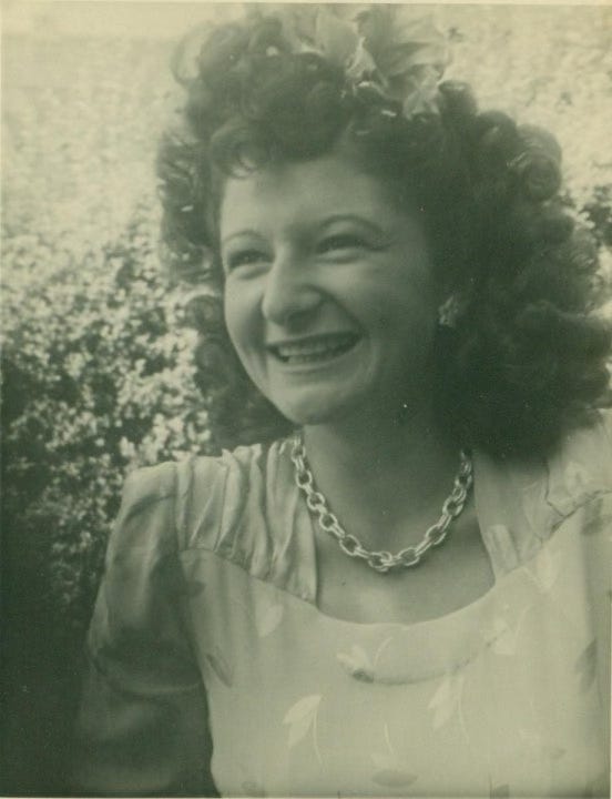 A faded photograph of a young white woman smiling big with a chain necklace and big earrings and wild, curly dark hair. 