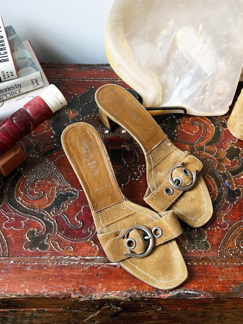 May include: A pair of brown suede heeled sandals with silver buckles. The sandals are on a wooden surface with a red and gold design.