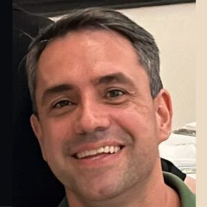 Homem sorridente, de cabelo curto e grisalho, vestindo uma camisa verde. O fundo é neutro, com alguns detalhes de objetos brancos ao fundo