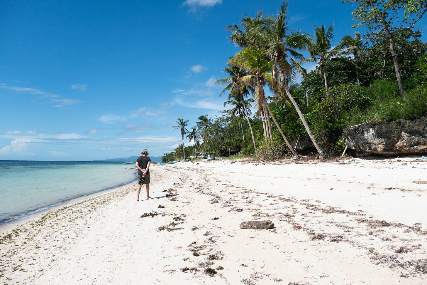 A woman walks along a sandy beach, the turquoise waters to her left, palm and coconut trees to her right, and mountains in the distance.