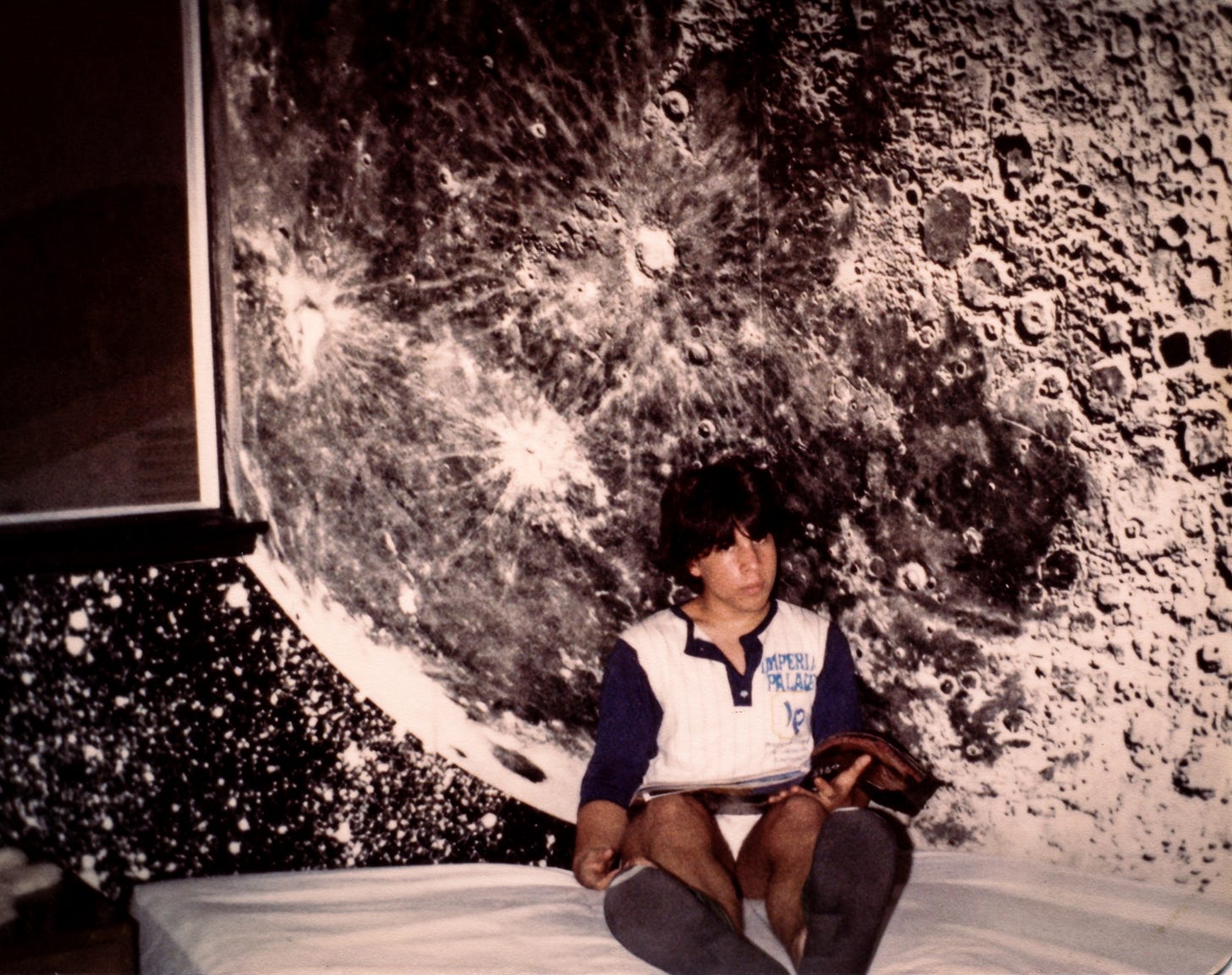 A teenaged boy in flip-flops sits on a bed reading an issue of Rolling Stone. The walls behind him are papered with a giant photo of the lunar surface.