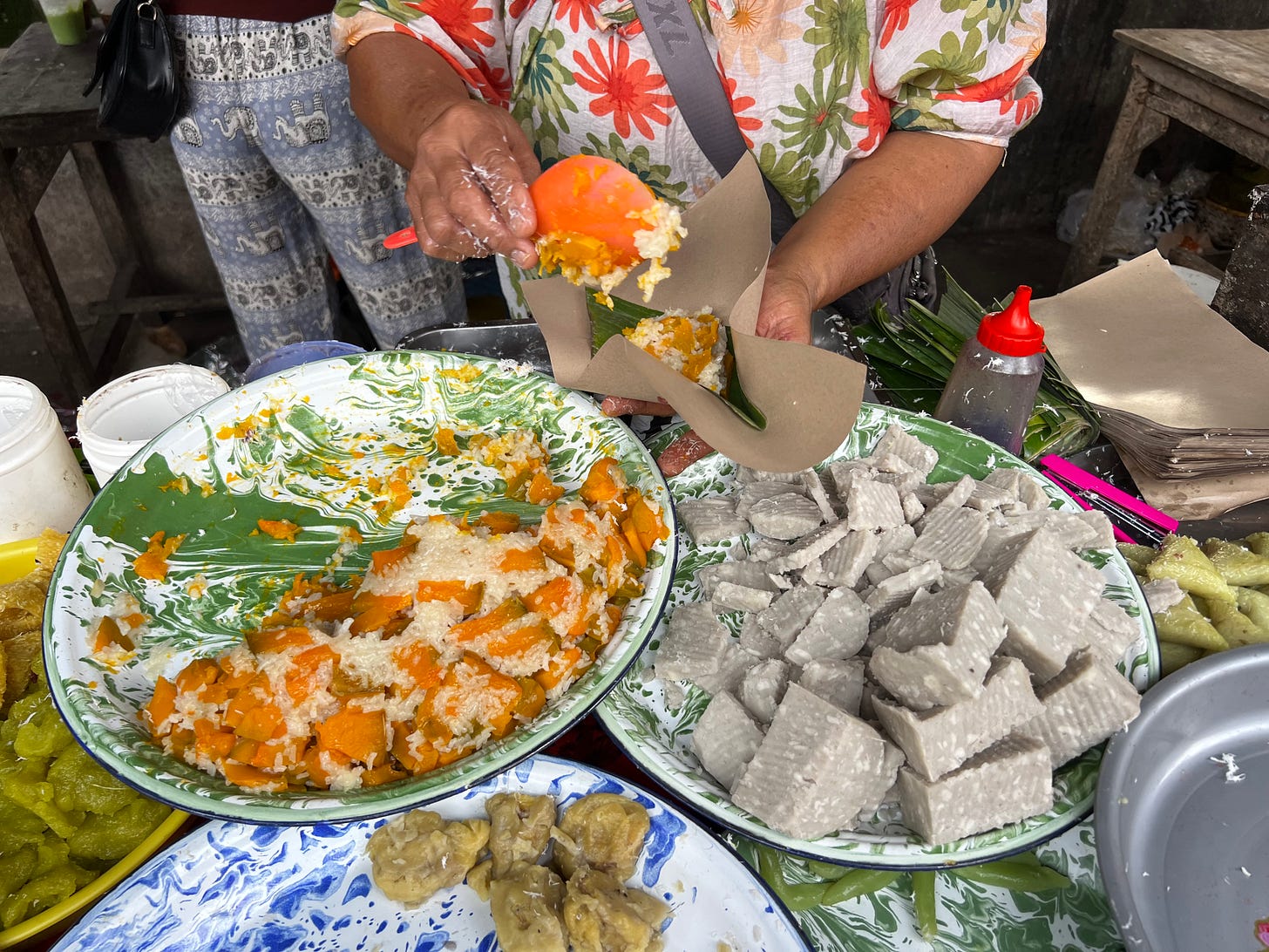 Hands spooning bright orange rice-and-coconut sweets into a cone of banana leaf and brown paper, over enamel platters of food. 